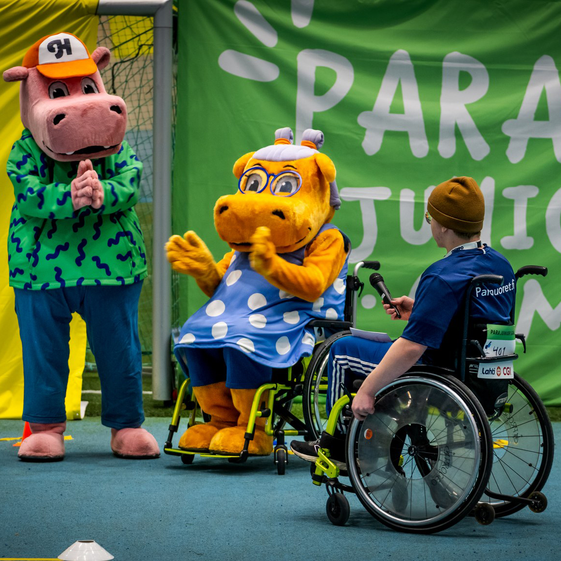 Pyörätuolia käyttävä Hippo-hahmo taputtaa ison vihreän Para Junior Games -lakanan edessä, seisova Hippo-hahmo vieressään. Hahmoja lähestyy pyörätuolia käyttävä poika mikrofonin kanssa.